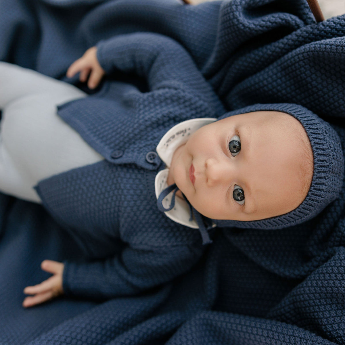 A baby lying on their back wrapped in a chunky navy blue blanket, wearing a matching navy knit cardigan and bonnet with a white collar.