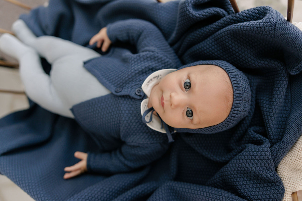 A baby lying on their back wrapped in a chunky navy blue blanket, wearing a matching navy knit cardigan and bonnet with a white collar.